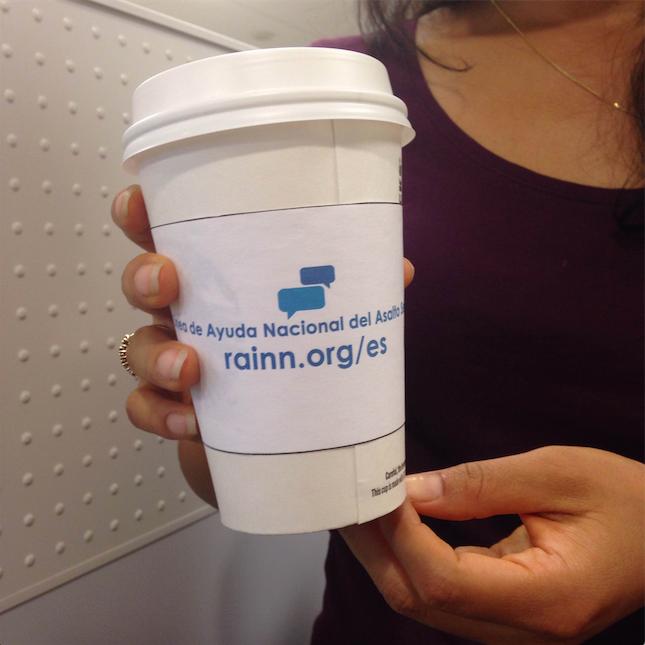 Person holds a coffee cup with a sleeve. On the sleeve is written "Linea de Ayuda Nacional del Asalto Sexual" and "rainn.org/es"